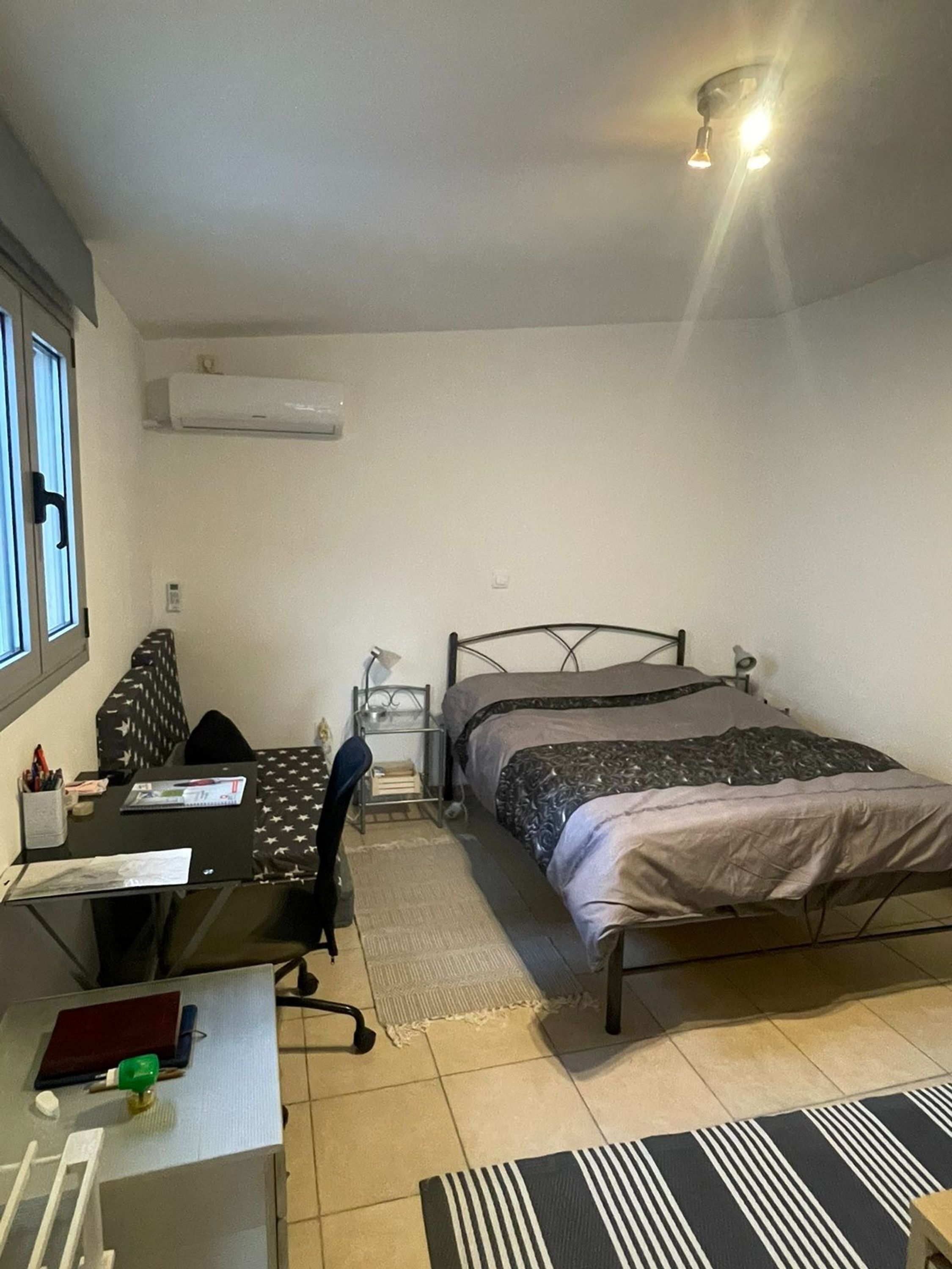 Een kleine, goed verlichte slaapkamer met een enkel bed, een bureau en een stoel. Aan één kant is er een raam en een airconditioningapparaat aan de muur.