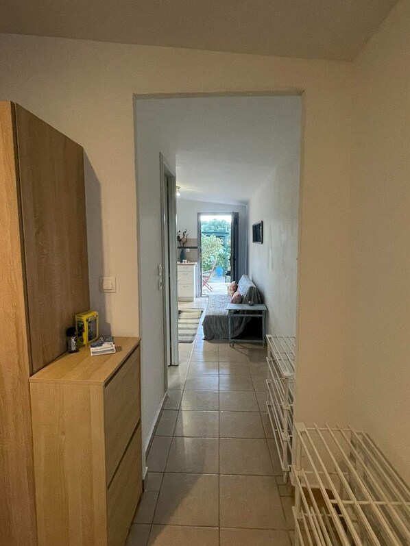A hallway leading to a bedroom with a bed, a small table, and a chair. There is a radiator on the right and a wooden cabinet on the left.