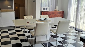 A dining area with a checkered floor, a white table, and four chairs. There is a microwave on a wooden cabinet, and a clock on the wall. - Casa Playa Direct (Dénia)