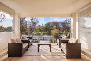 A balcony with wicker furniture and a glass table, overlooking a street and trees. - The Nepean | 100m to Beach, Pool & Fire Pit (McCrae)