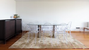 Ein zeitgemäßer Essbereich mit einem Glastisch, transparenten Stühlen, einem gemusterten Teppich und einem dunklen Schrank mit Pflanze darauf.