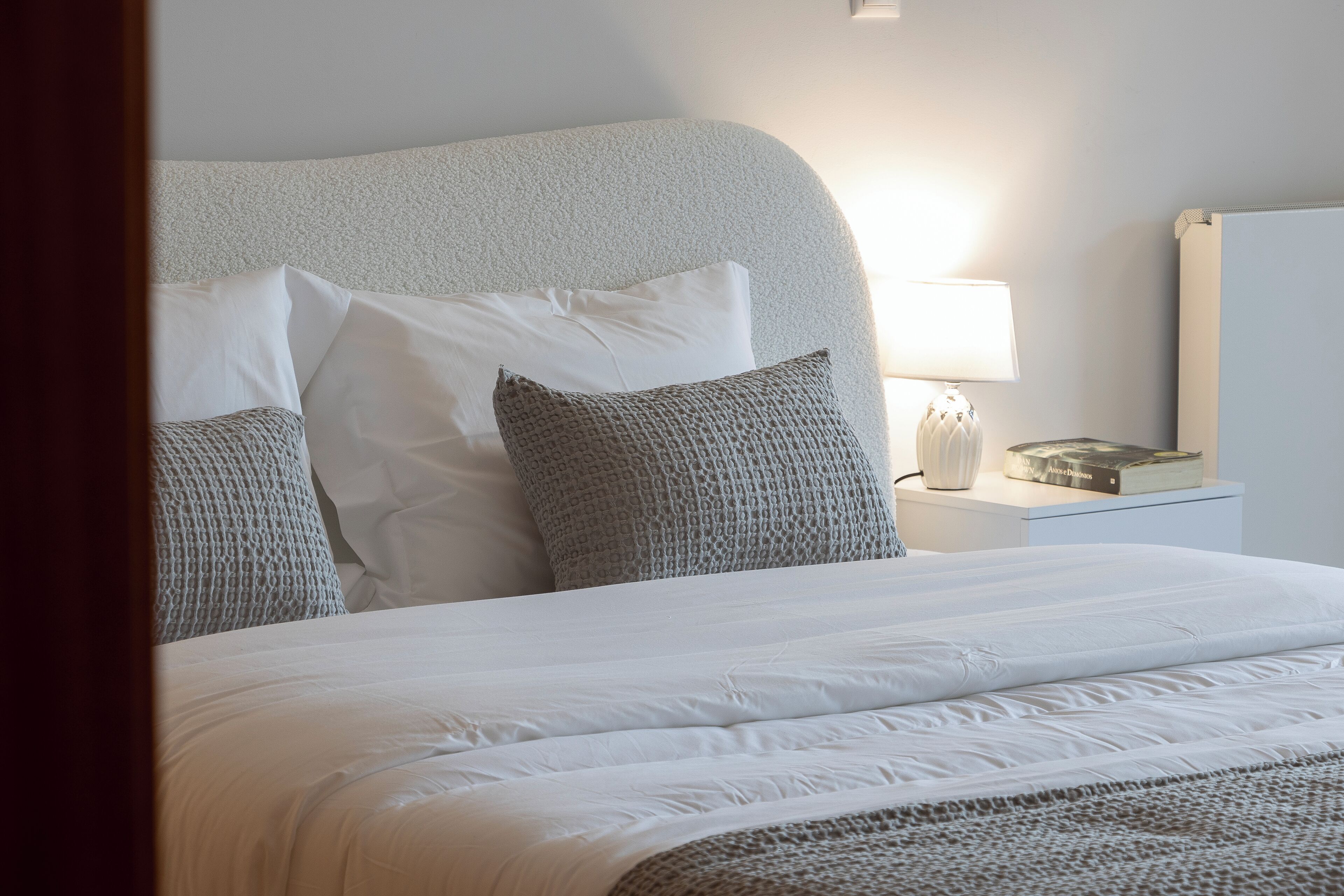 A neatly made bed with white linens and a grey knitted throw blanket. There is a bedside table with a lamp and a book.