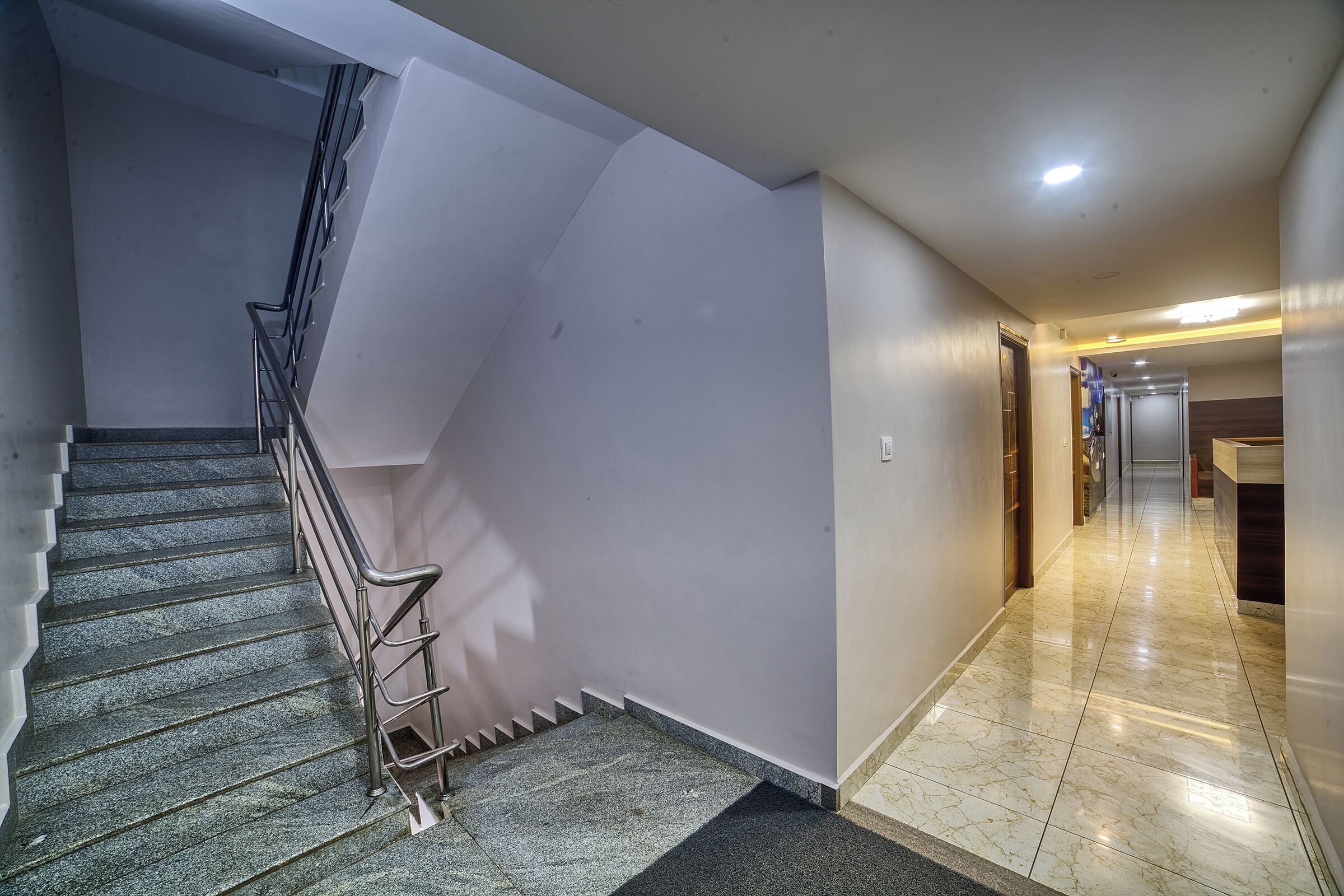 A modern hotel corridor with a marble floor, a staircase with metal railings, and a straight hallway leading to a reception area.