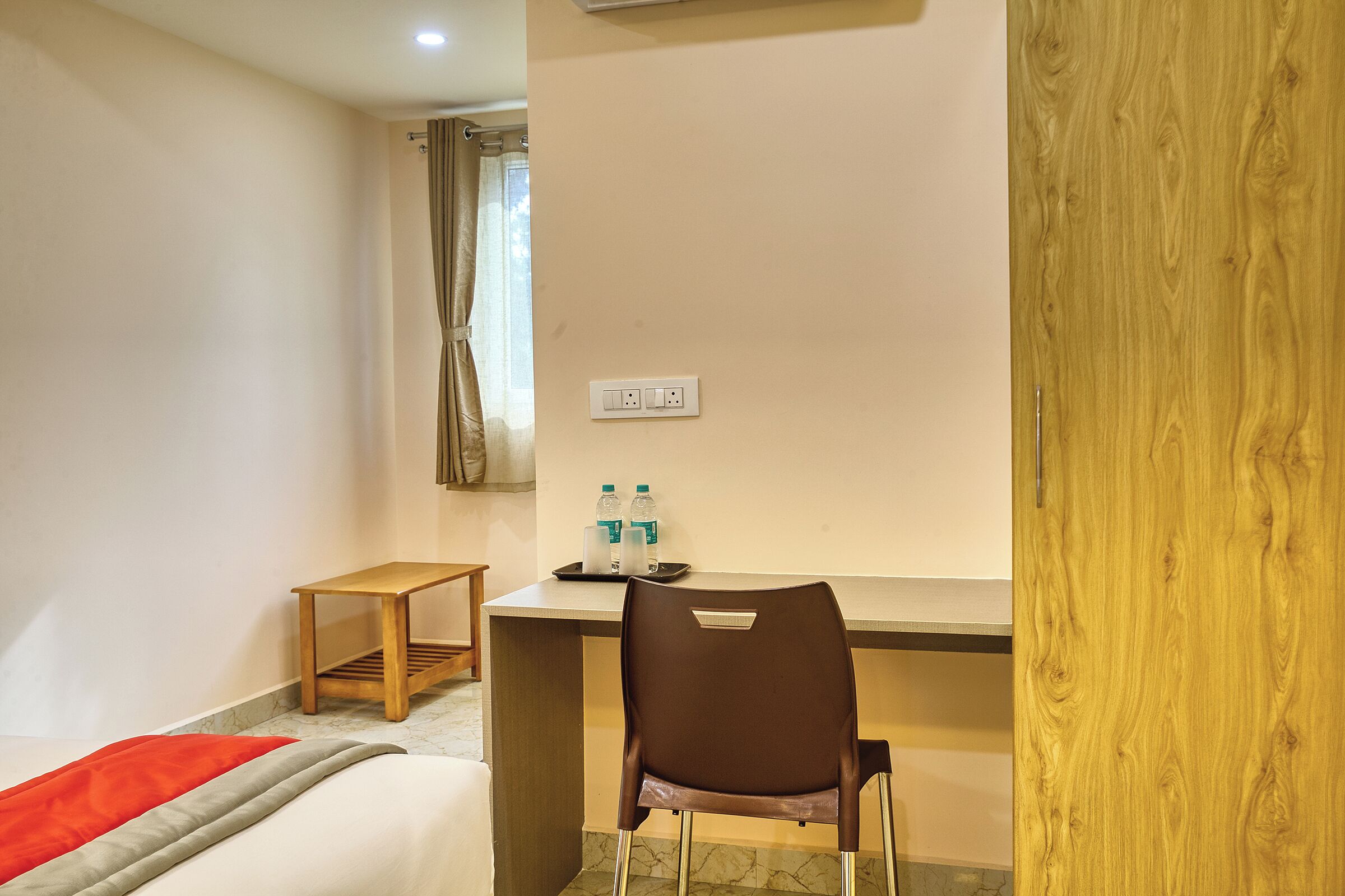 A hotel room with a bed, a desk, a chair, a small table, and a power outlet.