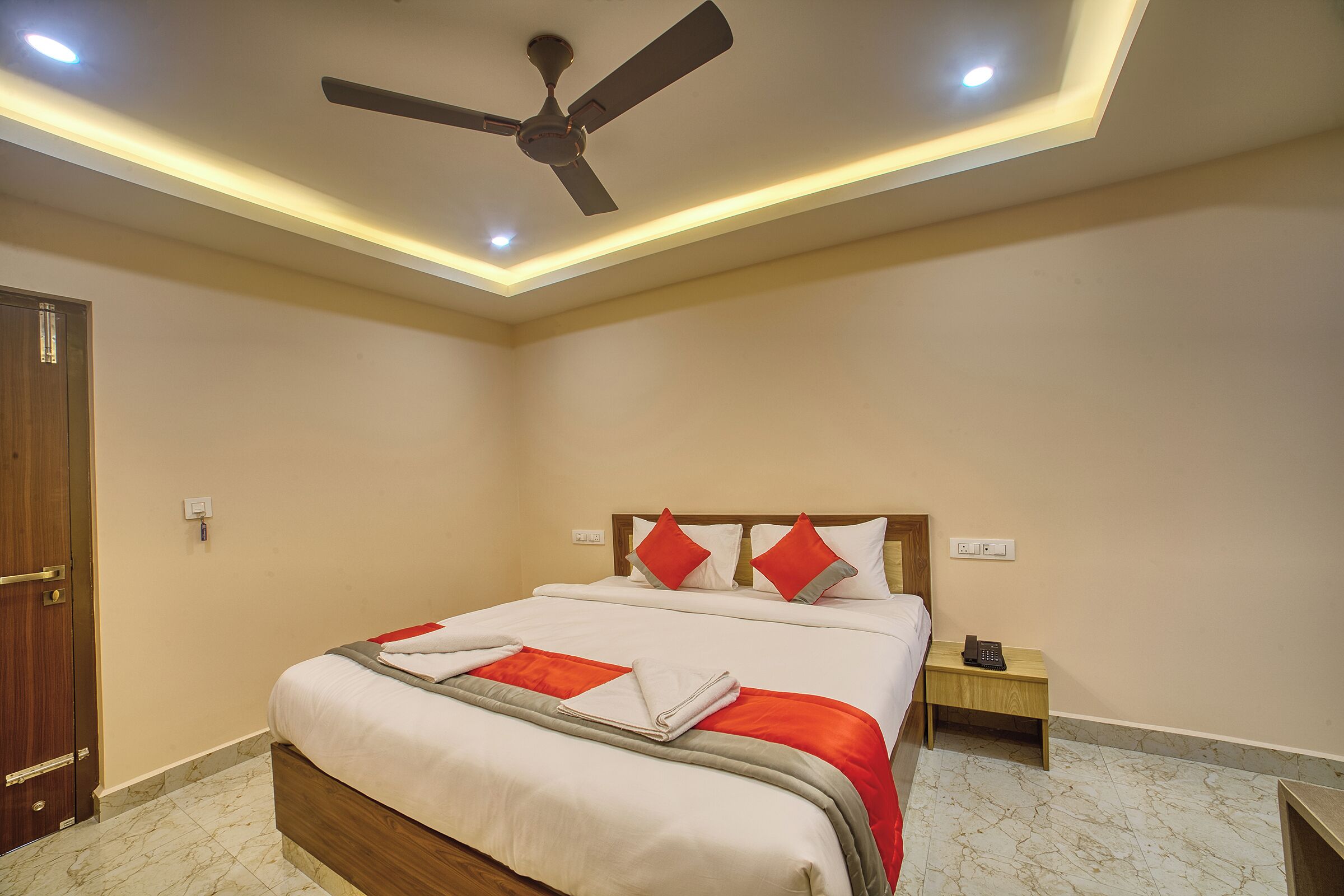 A hotel room with a bed, a ceiling fan, a bedside table with a phone, and a door.