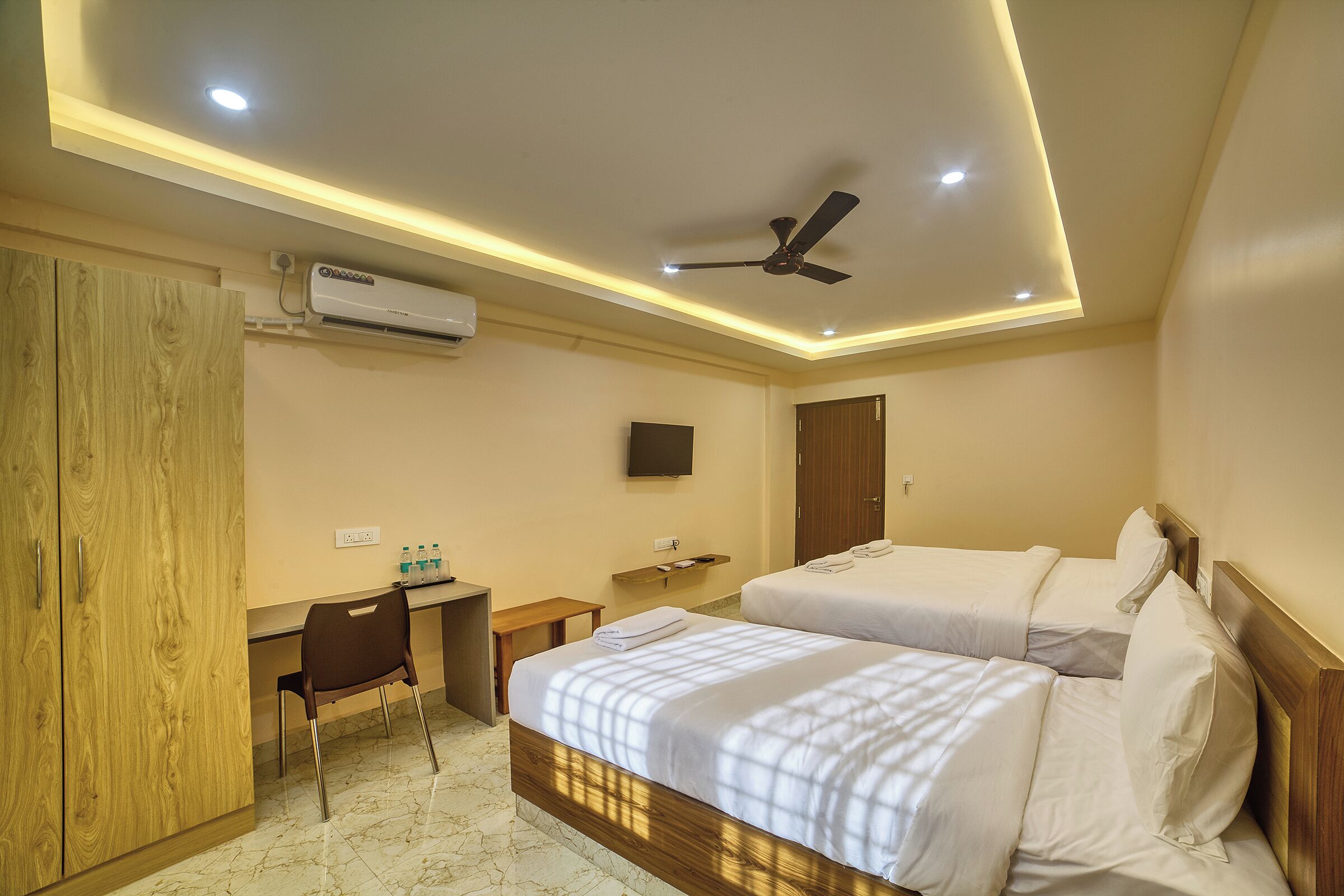 A hotel room with two beds, a desk, a chair, a TV, and an air conditioning unit.