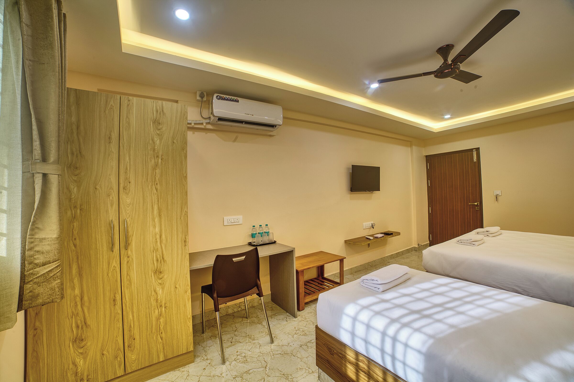 A hotel room with a desk, two beds, a ceiling fan, and an air conditioning unit.