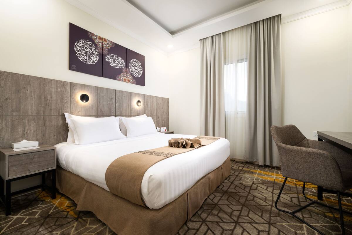 A hotel room with a large bed, a nightstand, a desk, and a chair. There are decorative wall art pieces above the bed and a window with curtains.
