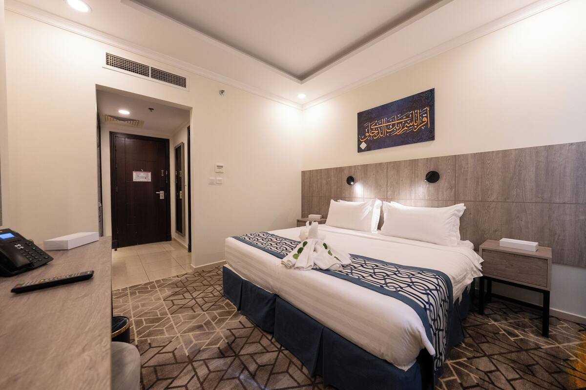 A hotel room with a bed, a bedside table, a nightstand, a remote control, and a wall-mounted artwork.