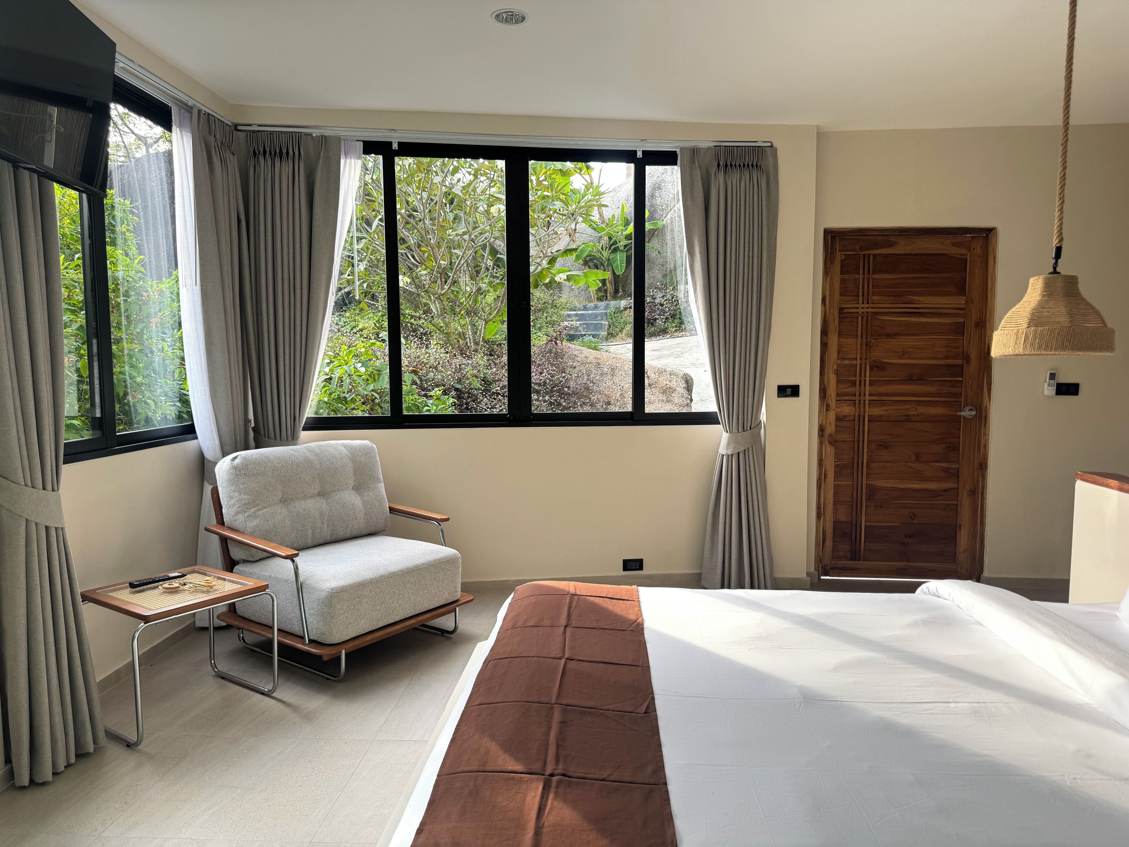 A bedroom with a bed, a chair, a small table, a window with curtains, and a wooden door.