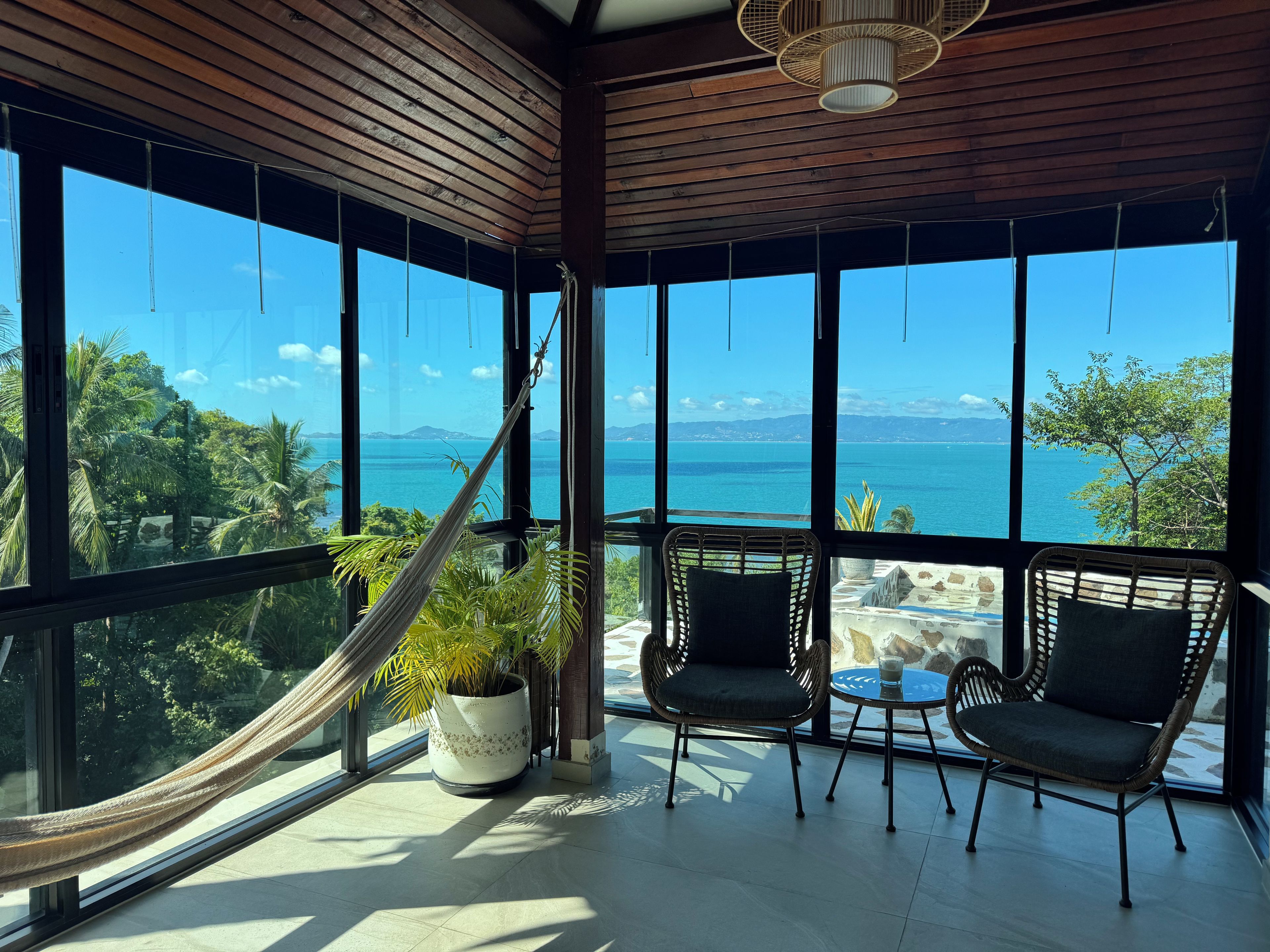 Una habitación con ventanas amplias, una hamaca, dos sillas de mimbre, una mesa y una planta en maceta.