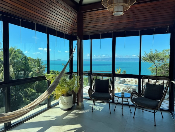 Una habitación con ventanas amplias, una hamaca, dos sillas de mimbre, una mesa y una planta en maceta.