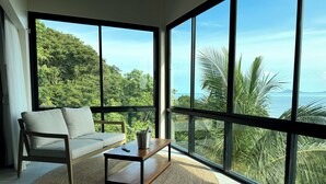 A modern living room with a sofa and a coffee table, large windows offering a view of lush greenery and the sea.