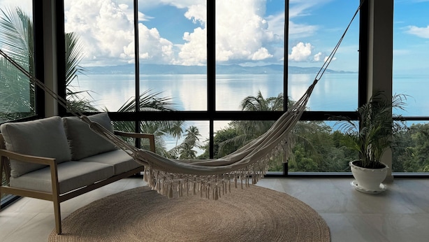 A hammock in a room with large windows overlooking a body of water and mountains.