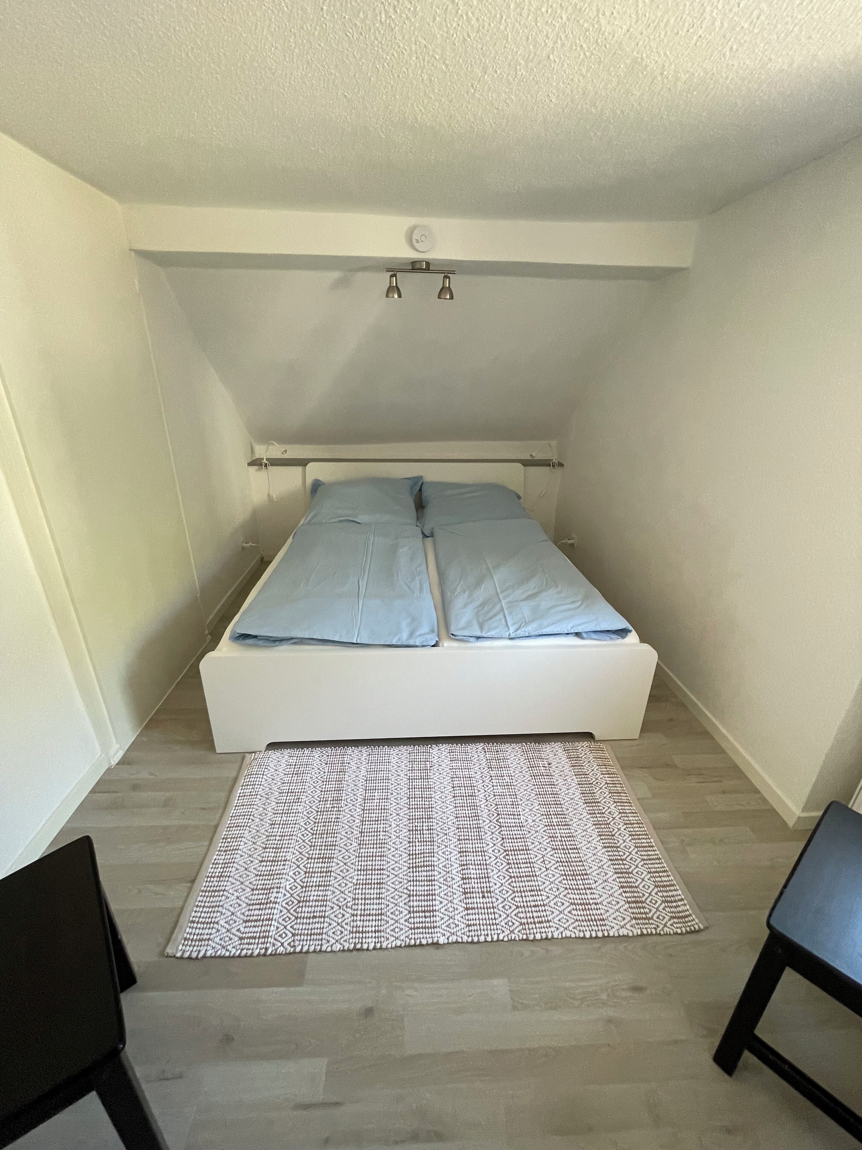 Ein kleines, helles Zimmer mit einem Einzelbett, einem gemusterten Teppich und zwei Stühlen.