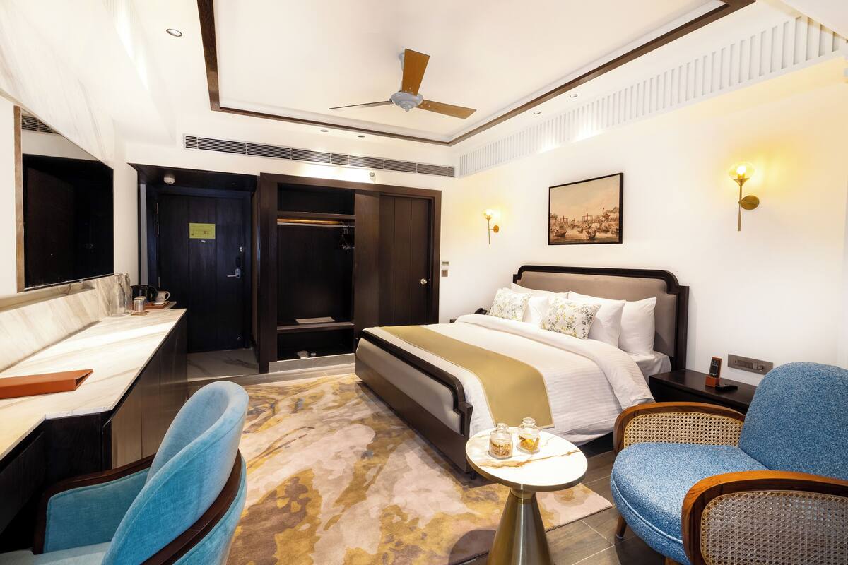 A hotel room with a bed, a desk, a chair, a small table, a ceiling fan, and a wall-mounted light.