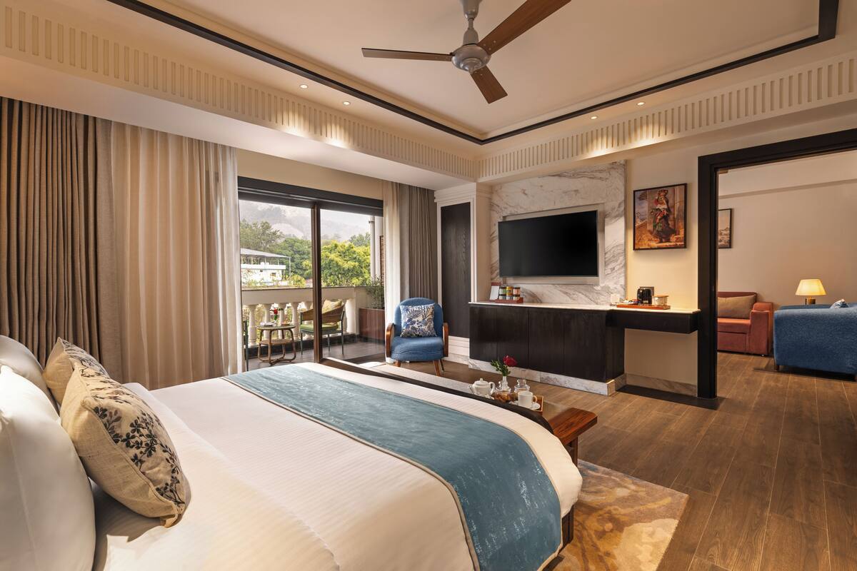 A spacious bedroom with a large bed, a desk, a TV, and a balcony with a view of the city.