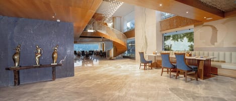 A spacious lobby with a marble floor, a wooden staircase, and a seating area with tables and chairs.