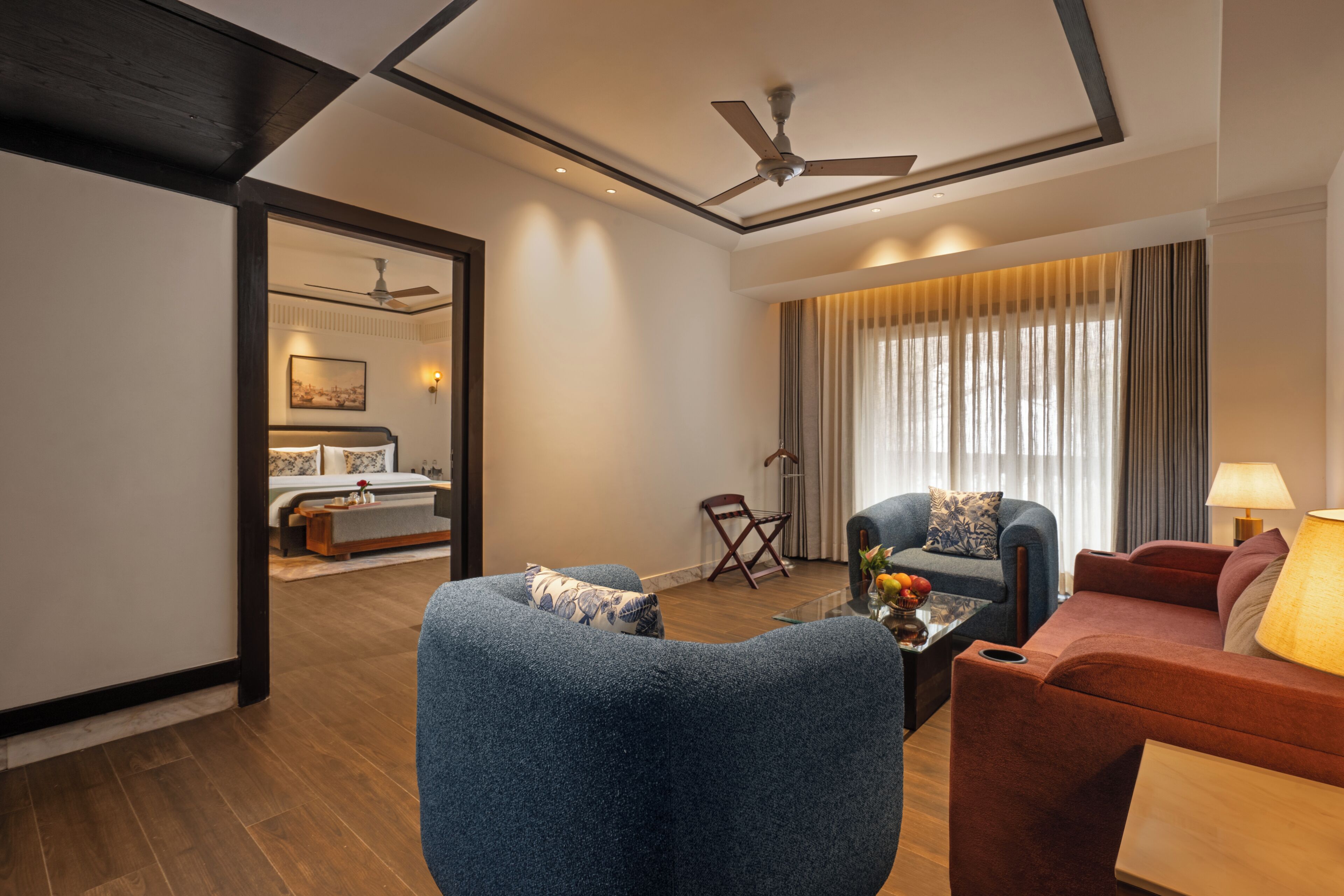 A modern hotel room with a large bed, a sitting area with a blue armchair and a red sofa, a ceiling fan, and a large window with curtains.