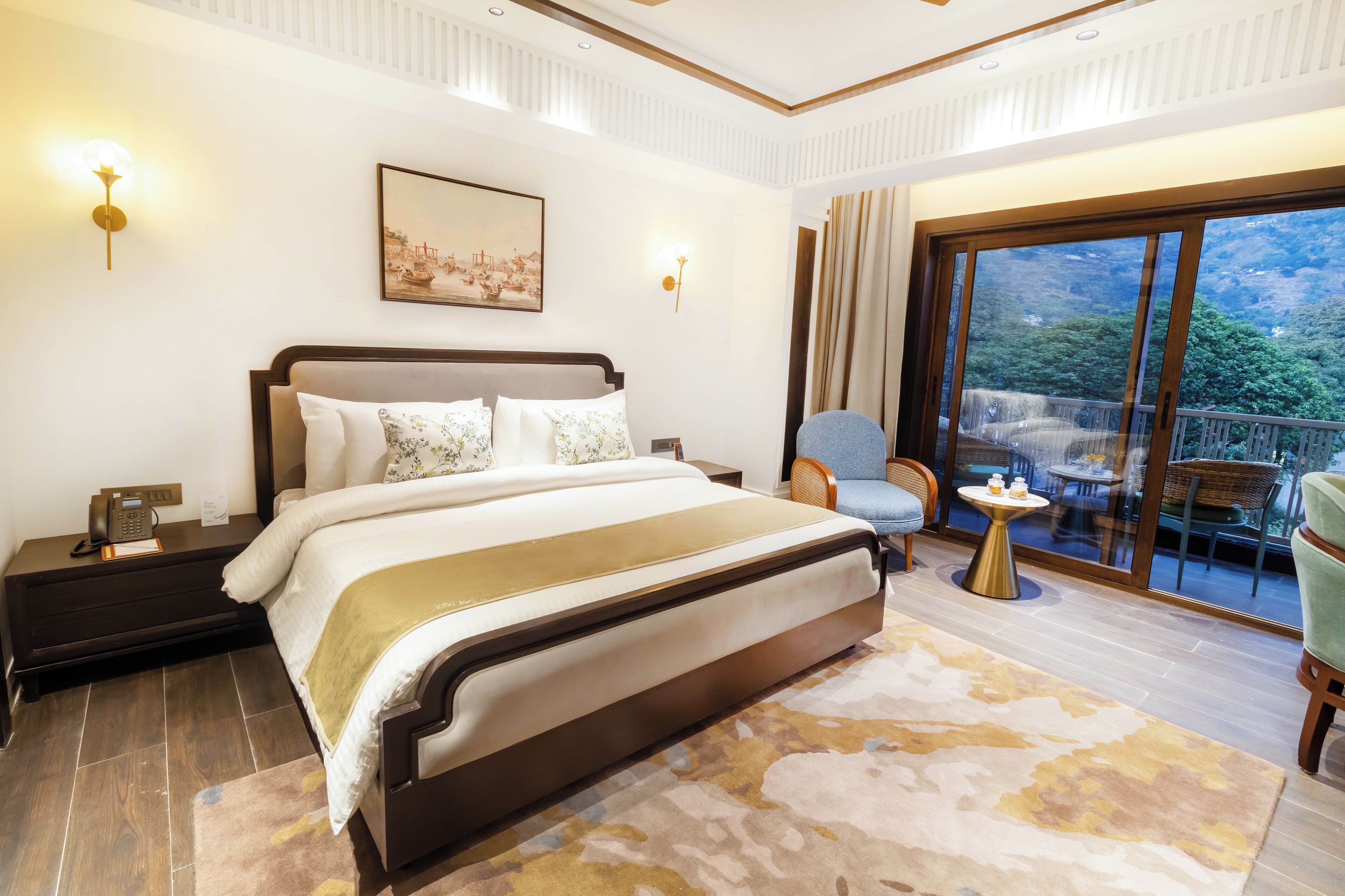 A bedroom with a large bed, a nightstand, a bedside table, a chair, a small round table, and a view of a balcony with outdoor furniture.