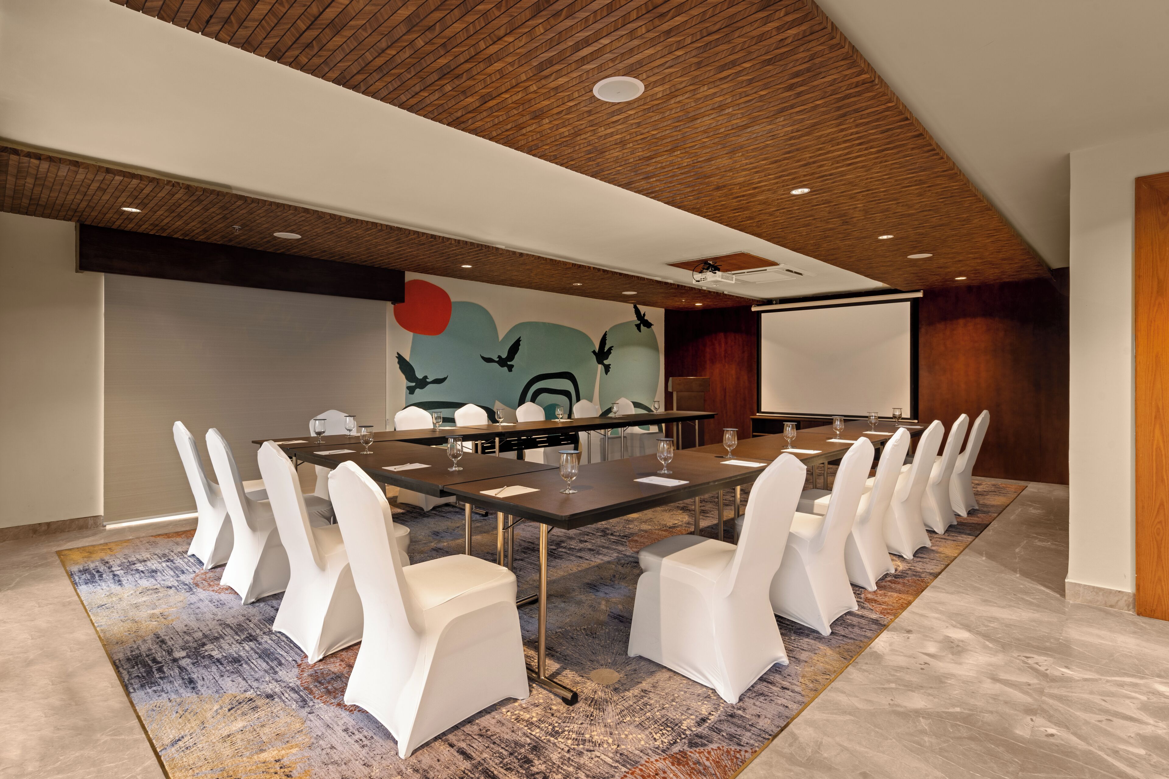 A conference room with a long table, white chairs, a projector screen, and a mural on the wall.
