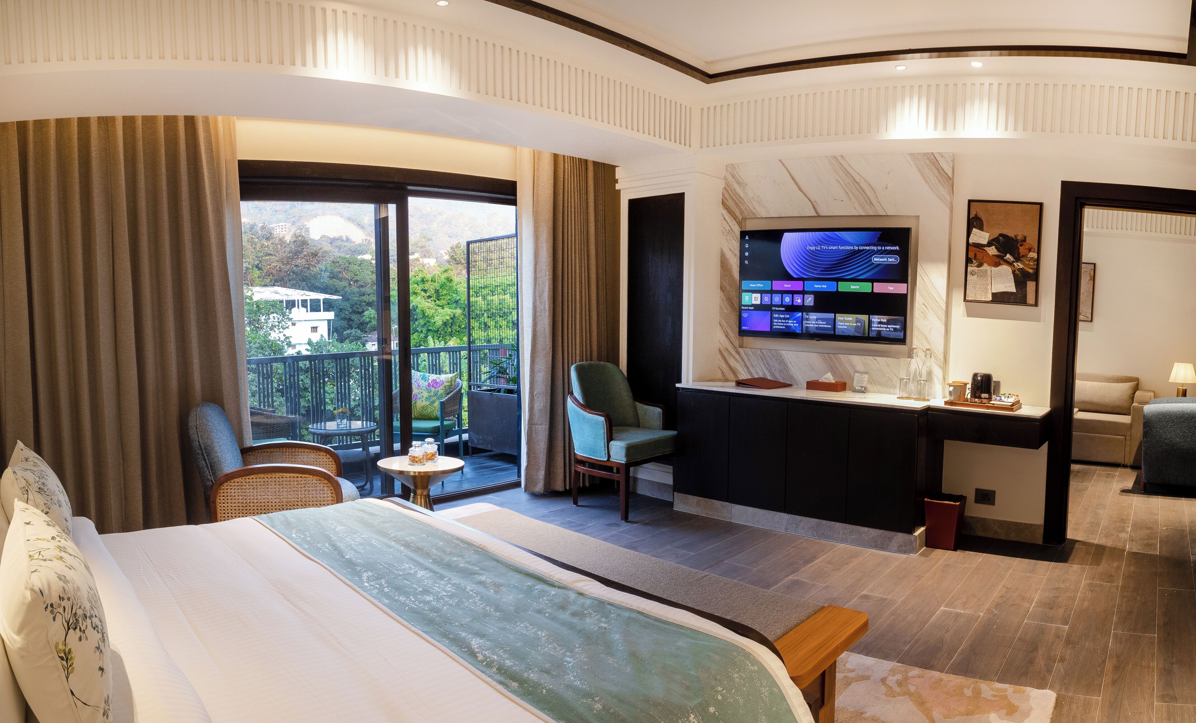 Une chambre d’hôtel comprenant un lit, un bureau avec un ordinateur, une chaise, un canapé et un balcon donnant sur un espace verdoyant.