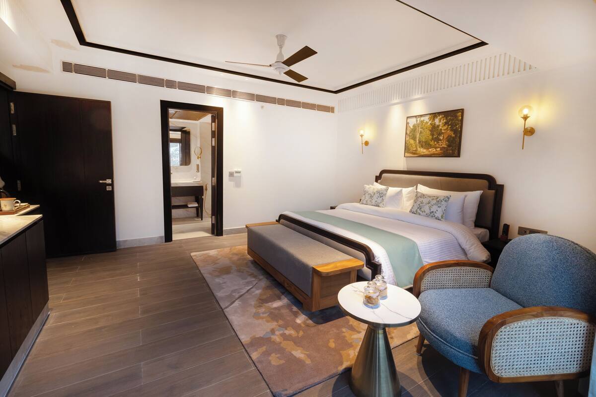 A modern hotel room with a large bed, a seating area with a bench and a chair, a small table with a lamp, and a ceiling fan.