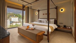 A four-poster bed with a canopy, a bedside table with a lamp, a nightstand, and a view of the cityscape through large windows. - Auradoon Dehradun - By Leisure Hotels (Dehradun)