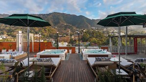 A rooftop terrace with hot tubs, lounge chairs, and umbrellas, overlooking a mountainous landscape. - Auradoon Dehradun - By Leisure Hotels (Dehradun)