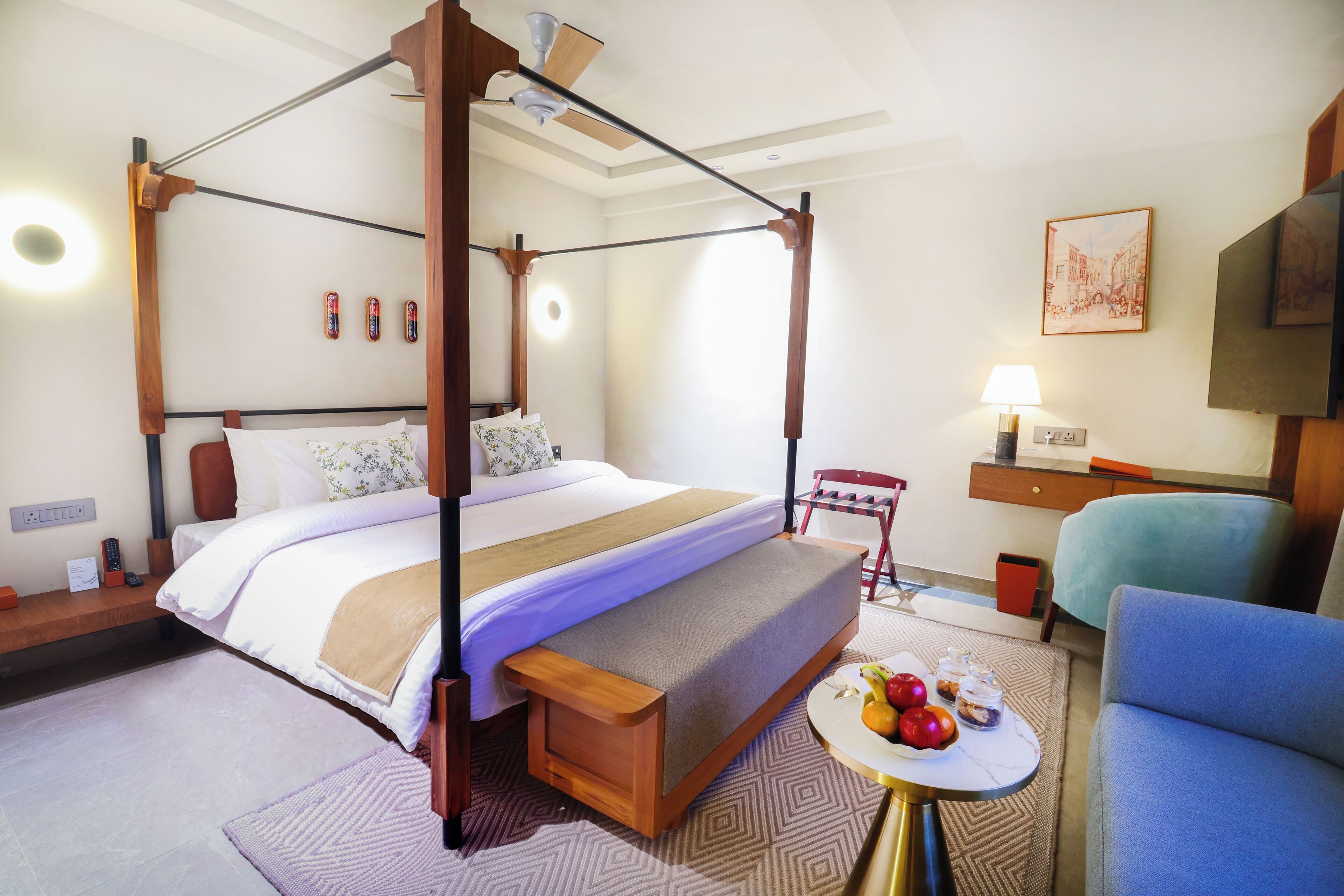 Een hemelbed met houten frame, een nachtkastje met lamp, een klein rond tafeltje met fruit en een vaas, en een blauwe fauteuil.