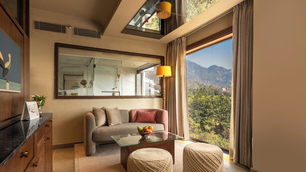 A modern hotel room with a sofa, a glass coffee table, two ottomans, a large mirror, and a view of mountains through the window. - Auradoon Dehradun - By Leisure Hotels (Dehradun)