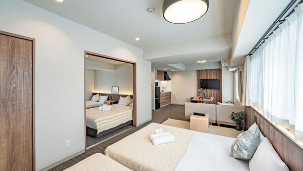 Une chambre d’hôtel moderne avec deux lits, un coin salon, une kitchenette et une grande fenêtre avec des rideaux.