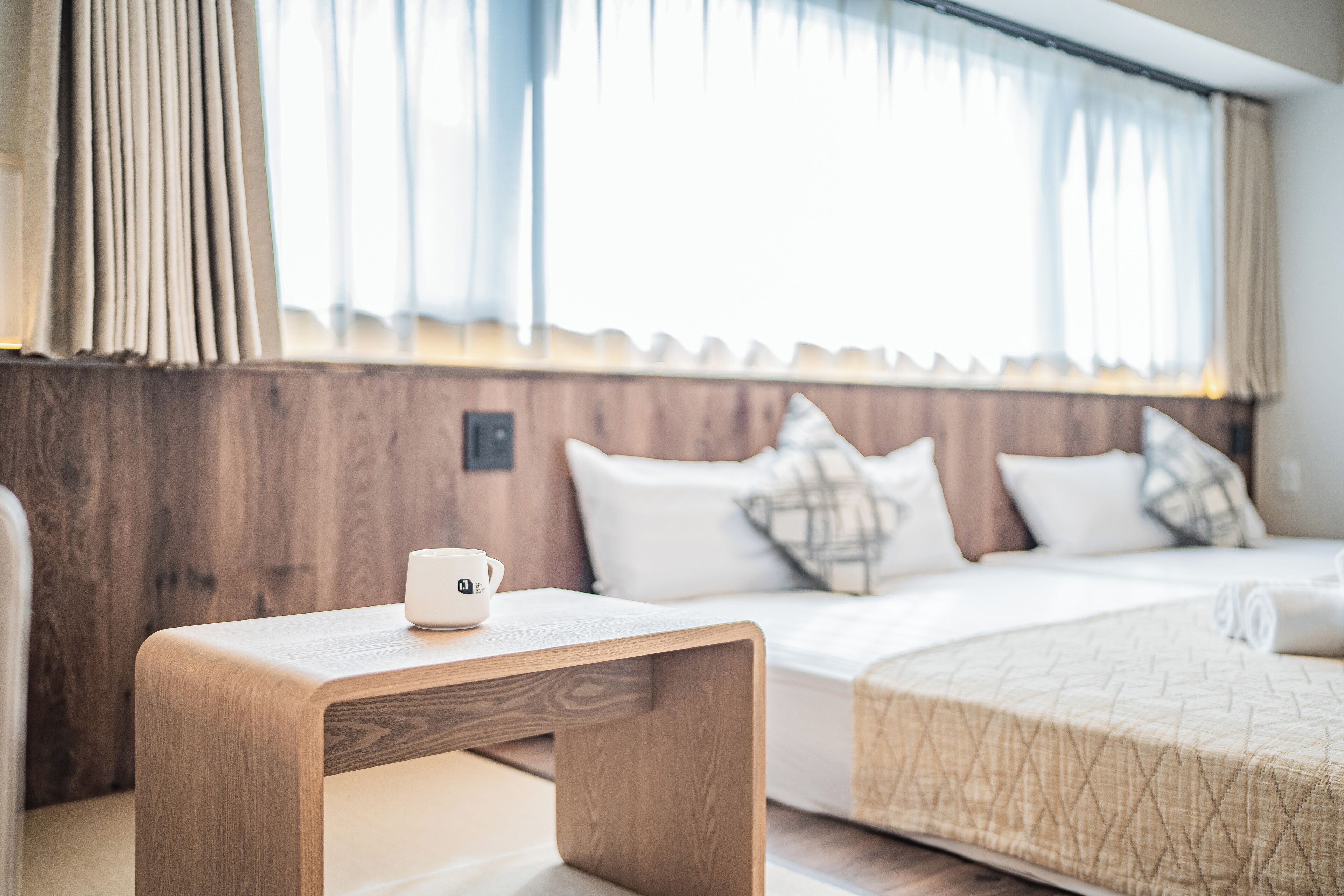 A hotel room with a wooden bedside table, a bed with a quilted bedspread, and a window with curtains.
