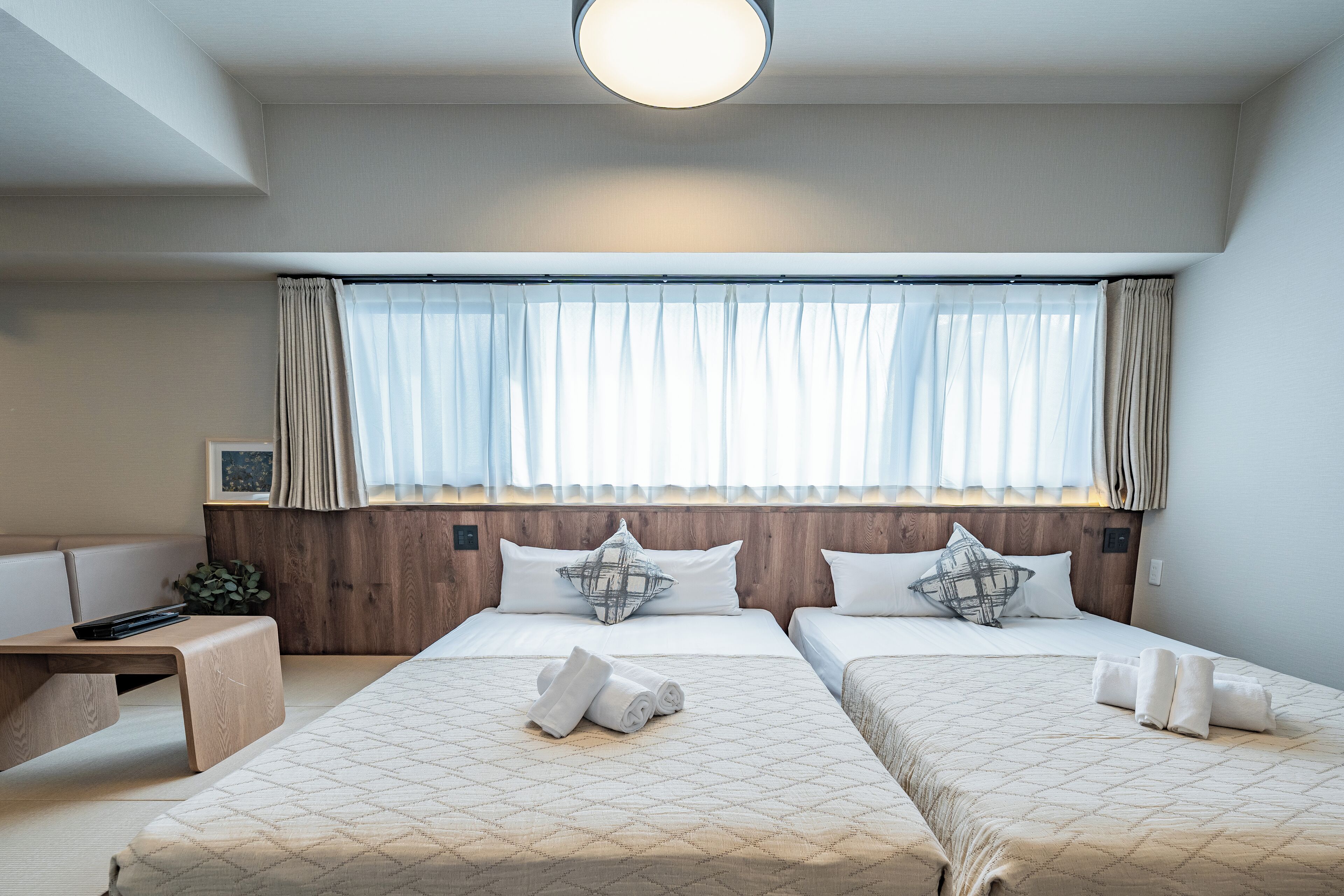 A hotel room with two beds, a bedside table, a small seating area, and a window with curtains.