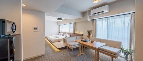 A modern hotel room with a kitchenette, a sofa bed, a dining table, and a large window with curtains.