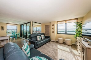 A modern living room with a black leather sofa, a dining area with green chairs, and large windows with a view of the water. - 2BR waterfront condo with balcony, pool, hot tub, and beachy views. (St. Petersburg)