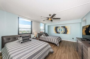 A modern hotel room with two beds, a ceiling fan, large windows, artwork, and a TV.