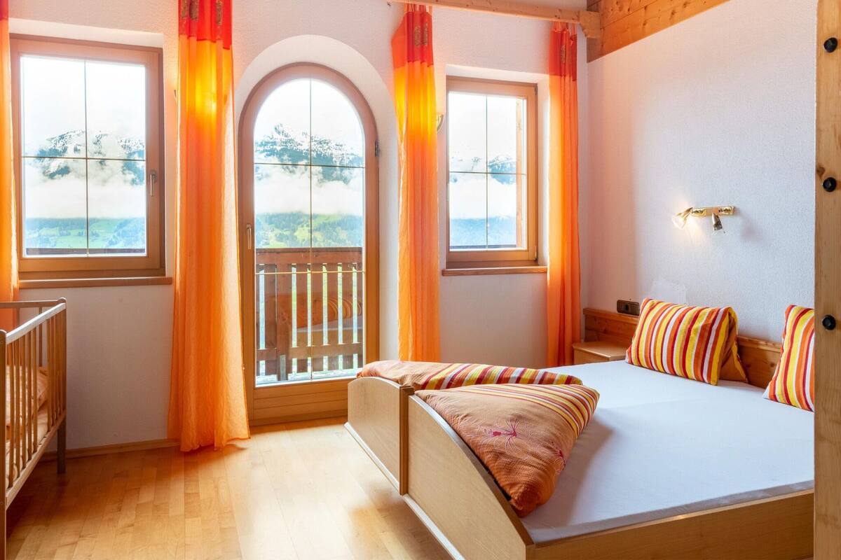 A bedroom with a wooden bed, two windows with curtains, a view of the outdoors, and a small balcony.
