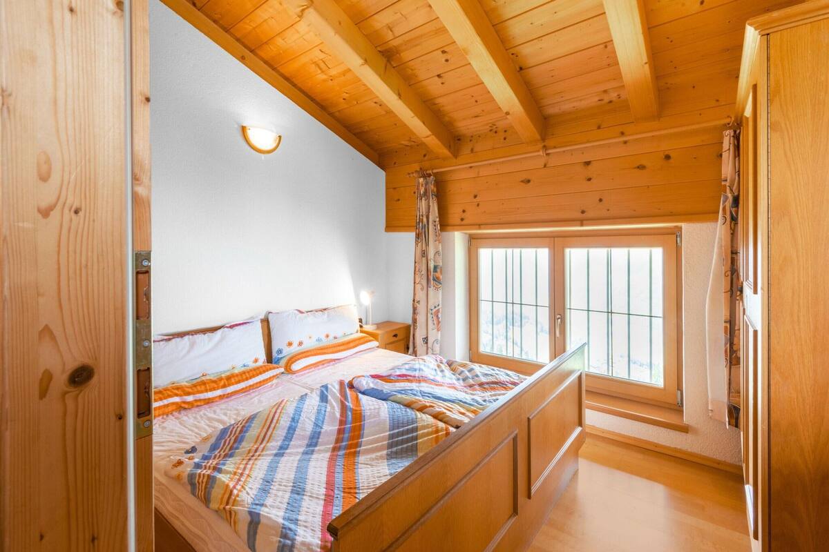A wooden cabin bedroom with a bed, a nightstand, a window with bars, and a wooden ceiling.