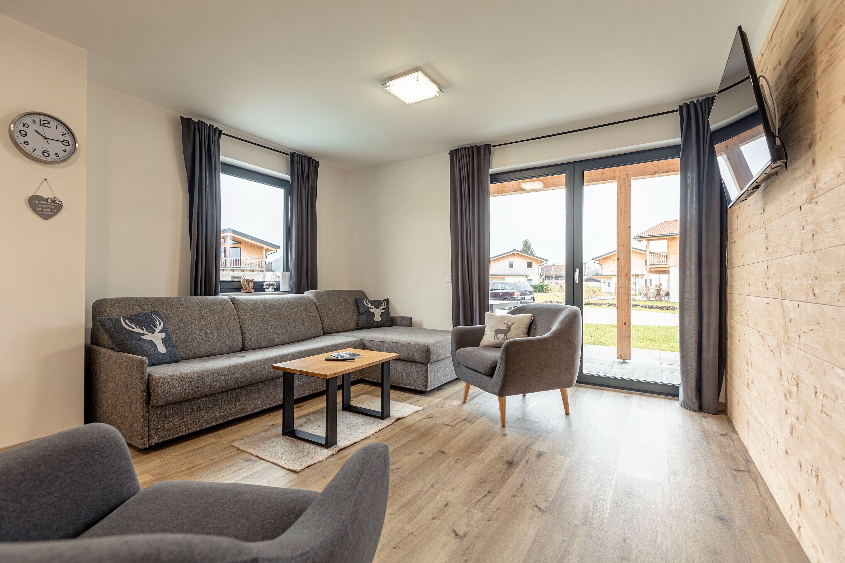A modern living room with a grey sofa, a wooden coffee table, and a grey armchair. There is a large window with curtains, a wall clock, and a flat-screen TV mounted on the wall.