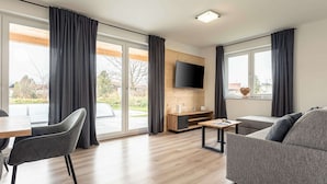 Een moderne woonkamer met een grijze bank, een eettafel met stoelen, een flatscreen televisie bevestigd aan een houten wand en grote ramen met gordijnen.