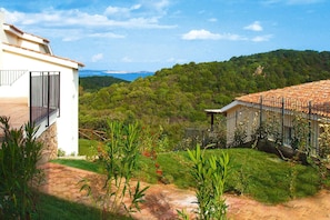 Un bâtiment blanc doté d’un balcon, un jardin avec des plantes et des fleurs, et une vue sur une colline boisée.
