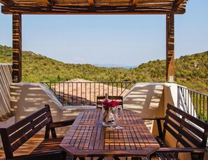 A wooden table with a vase of flowers and two benches under a pergola with a view of a hilly landscape. - Apartment in Golfo Aranci With Terrace (Arzachena)