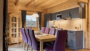 A wooden interior with a dining table and purple chairs, a kitchen with grey cabinets, and a window with curtains. - Chalets Almdorf Zillertal, Fugen (Fuegen)