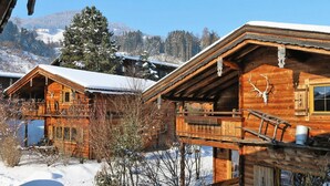 A wooden cabin with a balcony and a snow-covered roof, surrounded by a snowy landscape and mountains. - Chalets Almdorf Zillertal, Fugen (Fuegen)