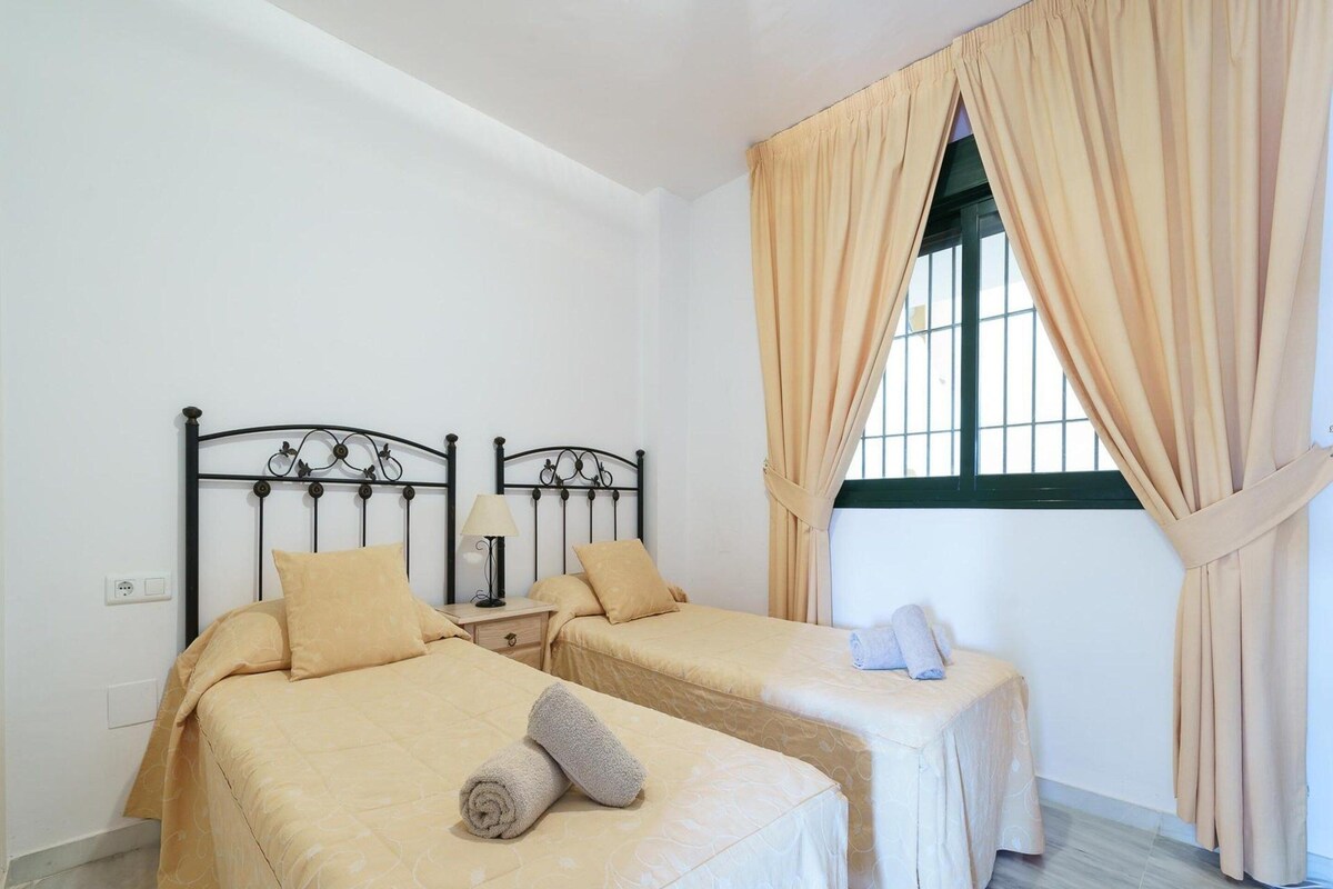 A room with two beds, a nightstand, a lamp, and a window with curtains.