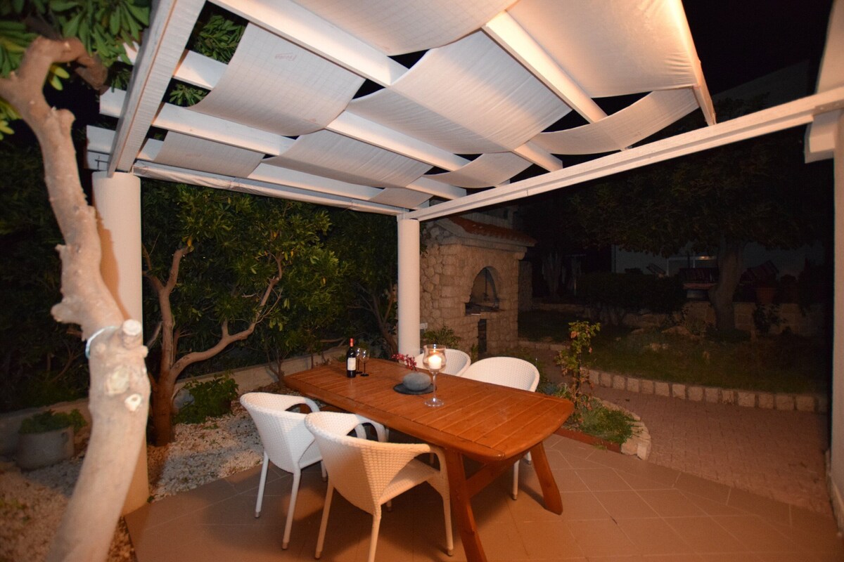 An outdoor patio with a wooden table and white chairs, a stone wall, and a pergola structure.