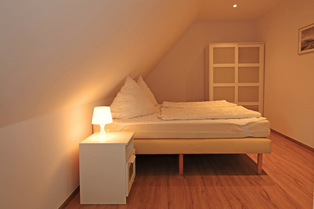 A single bed in a room with a sloped ceiling, a bedside table with a lamp, and a window with a view of the outside.