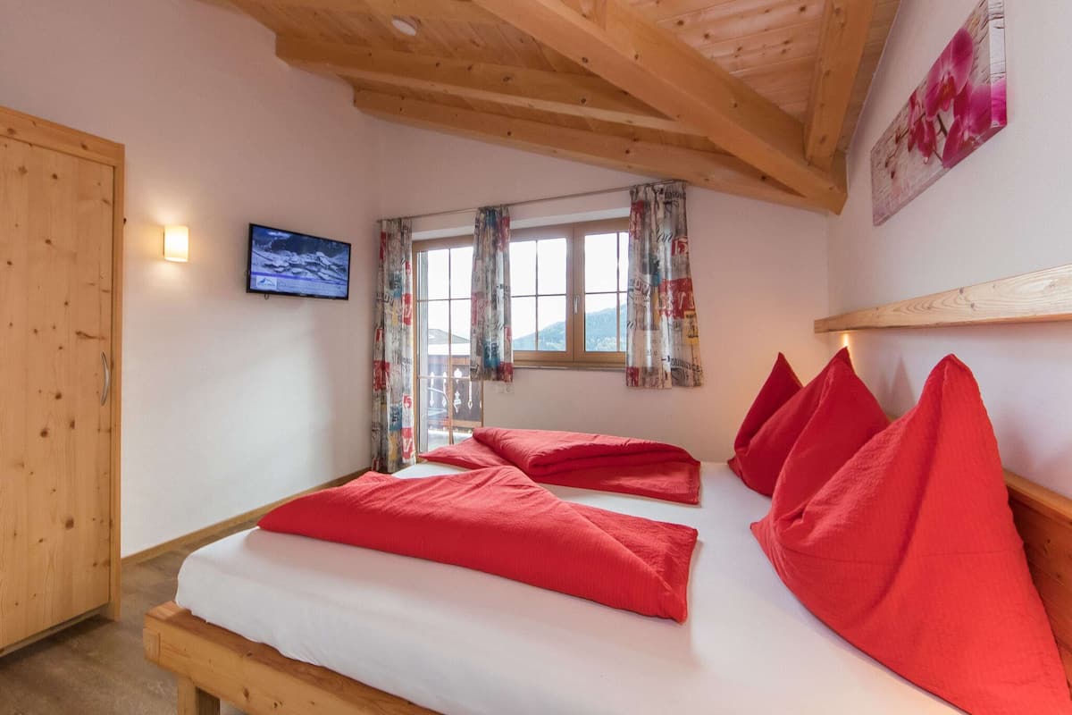 Un dormitorio con techo de madera, una cama con almohadas rojas, un televisor en la pared y una ventana con cortinas estampadas.