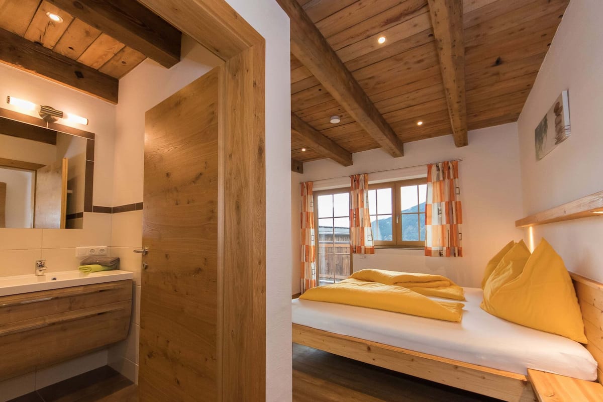 Ein Schlafzimmer mit Holzrahmen, einem Bett, einem Fenster mit Vorhängen sowie einem Badezimmer mit Waschbecken und Spiegel.